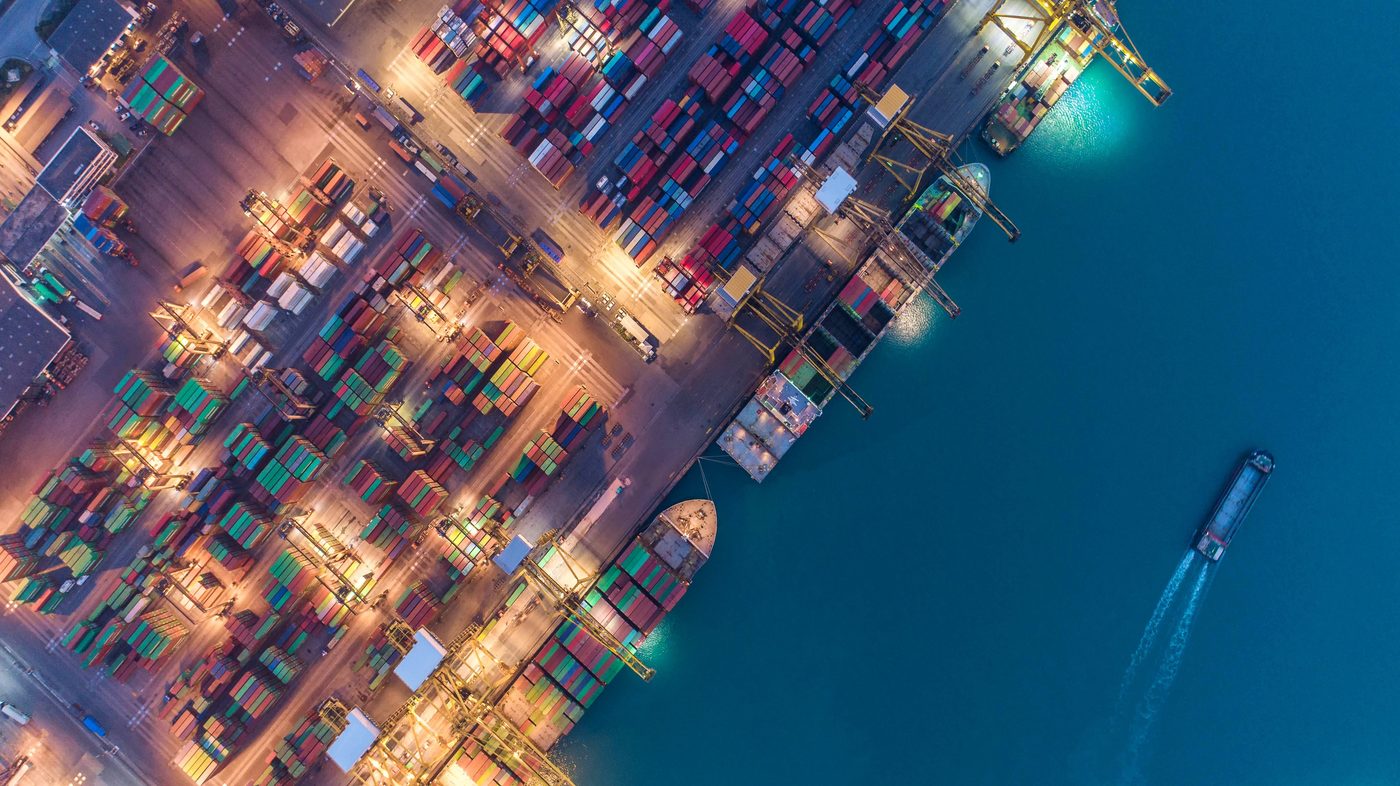 Aerial view of a busy container terminal at twilight, with illuminated docks and a vessel cruising past